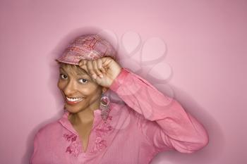 Portrait of smiling African- American young adult woman on pink background tipping her hat.
