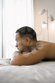 Profile of shirtless Asian young adult man lying on bed resting head on arms looking out window.