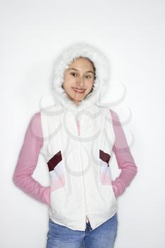 Portrait of Asian-American teen girl with hands in coat pockets and furry hood on head smiling against white background.