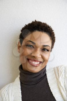 Close up head and shoulder of African- American woman standing against white wall smiling looking at viewer.