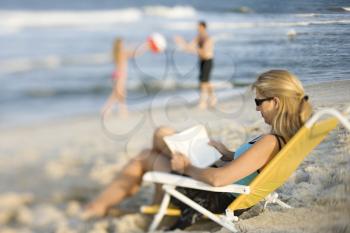 Royalty Free Photo of a Mother Reading in a Lounge Chair on a Beach While Husband and Daughter Play Ball in the Background