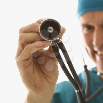 Mid-adult Caucasian male doctor in scrubs holding a stethoscope out towards viewer.