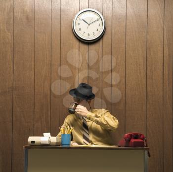 Royalty Free Photo of a Retro Businessman Sitting at a Desk Wearing a Fedora and Drinking Coffee