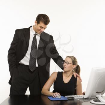 Caucasian mid-adult man touching shoulder of woman sitting at computer who feels uncomfortable.