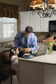 Caucasian father in suit using laptop computer with daughter eating breakfast in kitchen.