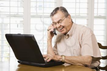 Royalty Free Photo of an Older Man Typing on a Laptop and Talking on a Cellphone