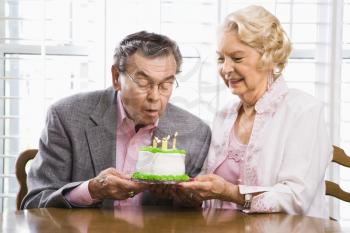 Mature Caucasian man blowing candles out on birthday cake while Mature Caucasian woman watches.