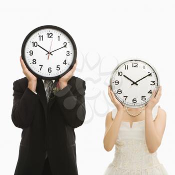 Portrait of Caucasian groom and Asian bride with clocks covering their faces. 
