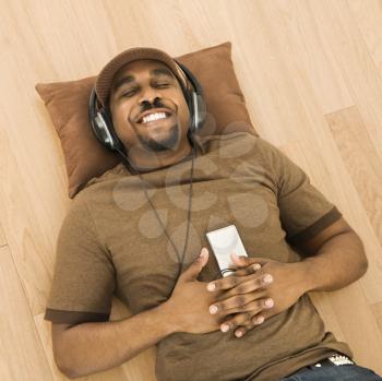 African-American mid-adult man wearing headphones and listening to mp3 player while lying on back on floor.