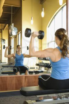 Woman sitting on a weightlifting bench. She is looking in a mirror while lifting dumbbells. Vertical shot.