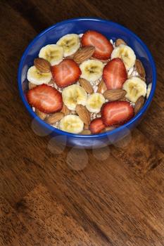 A blue bowl filled with bananas, strawberries, almonds and cereal. It sits on a wood table. Vertical shot.
