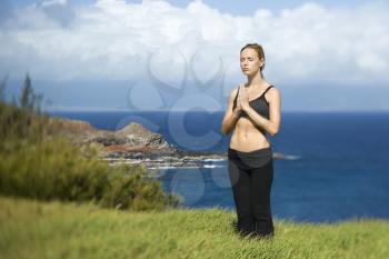 Attractive young woman stands with eyes closed in meditation in a green field overlooking the coast. Horizontal shot.