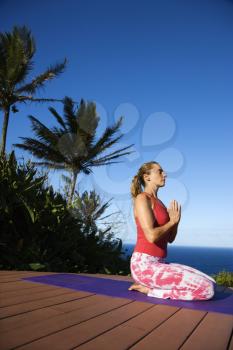 Attractive young woman in red sits on an exercise mat doing yoga with the ocean in the background. Vertical shot.