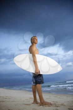 Young male surfer standing on beach with surfboard looking out at ocean.