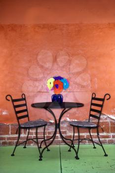 Outside patio table and chairs against a colorful wall.  There is a bouquet of paper flowers on the table. Vertical shot.