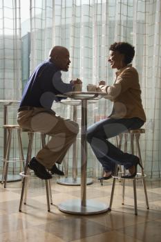 An African-American man and woman enjoy each other's company over a cup of coffee.  They are seated at a small cafe table on stools. Vertical shot.