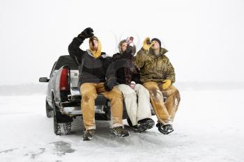 Two young men and a young woman drinking beer while sitting on the tailgate of a truck in a winter environment. Horizontal shot.