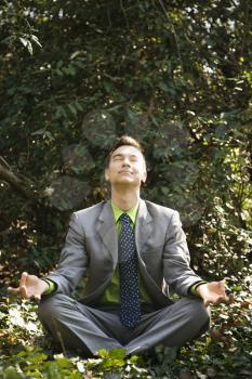 Young businessman sits in a lotus position meditating in the woods with closed eyes and a smile.