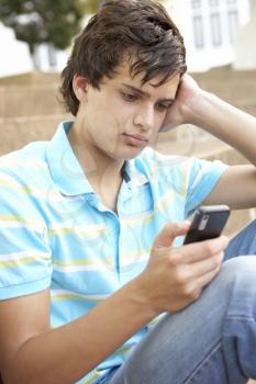 Unhappy Male Teenage Student Sitting Outside On College Steps Using Mobile Phone