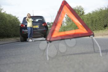 Female Driver Broken Down On Country Road With Hazard Warning Sign In Foreground