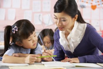 Teacher Helping Student Working At Desk In Chinese School Classroom