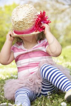 Young Girl In Summer Dress Sitting In Field Wearing Straw Hat