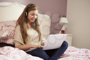 Teenage Girl Lying On Bed Using Laptop Wearing Headphones