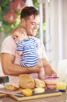 Father Holding Baby And Making Snack Whilst Using Laptop