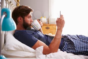 Man Eating Breakfast In Bed Whilst Using Mobile Phone