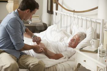 Doctor Taking Pulse Of Senior Male Patient In Bed At Home