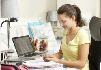 Teenage Girl Studying At Desk In Bedroom Using Mobile Phone