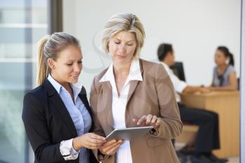 Two Female Executives Looking At Tablet Computer With Office Meeting In Background