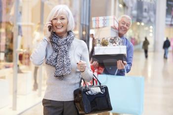 Senior Woman Shopping In Mall As Husband Carries Boxes