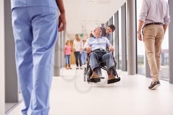 Nurse Pushing Senior Patient In Wheelchair Along Corridor