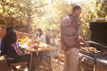 Man barbecues for friends at a table on a deck in a forest