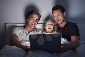 Young girl in bed watching laptop with her male gay parents