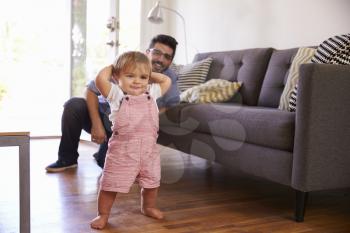 Father Watching Baby Daughter Take First Steps At Home