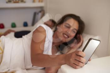 Woman in bed uses smartphone, partner sleeps, focus on phone