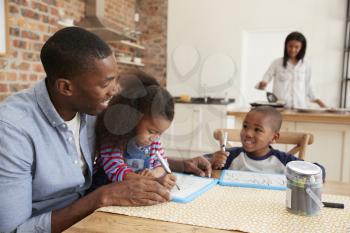 Father And Children Drawing At Table As Mother Prepares Meal