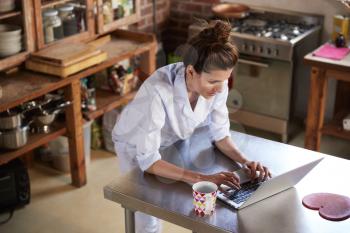 Woman in pyjamas stands using laptop in kitchen, high angle