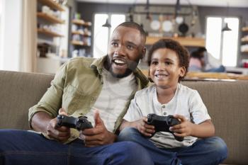 Father And Son Sitting On Sofa In Lounge Playing Video Game