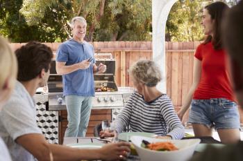 Family at a table outdoors turn to dad standing by barbecue