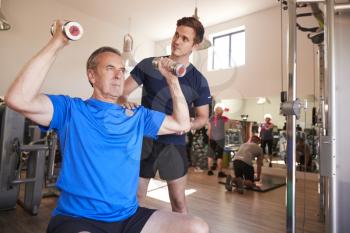 Senior Man Exercising With Weights Being Encouraged By Personal Trainer In Gym
