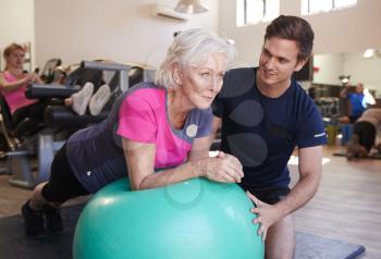 Senior Woman Exercising On Swiss Ball Being Encouraged By Personal Trainer In Gym