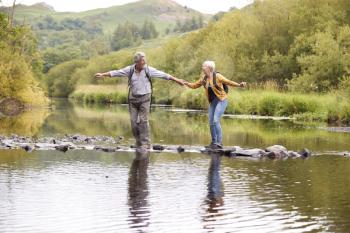 Senior Couple Crossing River Whilst Hiking In UK Lake District