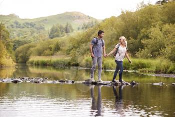 Couple Crossing River Whilst Hiking In UK Lake District