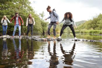 Multi ethnic group of five young adult friends laughing as they balance on rocks to cross a stream during a hike