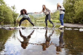 Three young adult women hold hands helping each other while carefully crossing a stream on stones during a hike