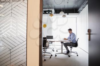 Businessman Working On Laptop At Boardroom Table Viewed Through Meeting Room Door