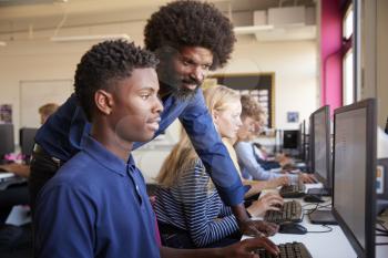 Teacher Helping Teenage Male High School Student Working In Computer Class